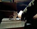 He finds a pair of horn-rimmed glasses on a table.